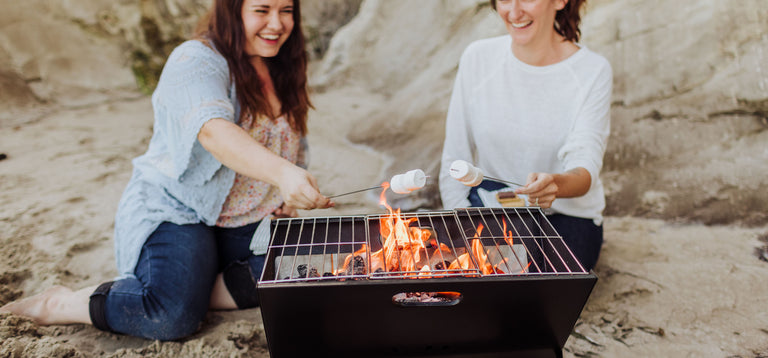 two woman making s'mores at the beach with portable folding charcoal grill